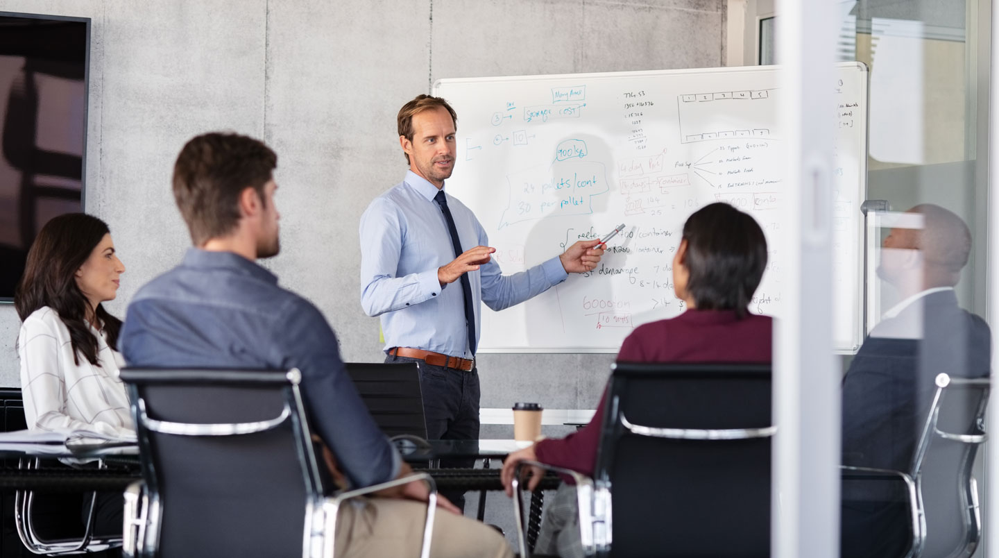 Formateur écrivant sur un tableau blanc émaillé dans une salle de formation professionnelle lumineuse, marqueur en main, groupe de participants en arrière-plan